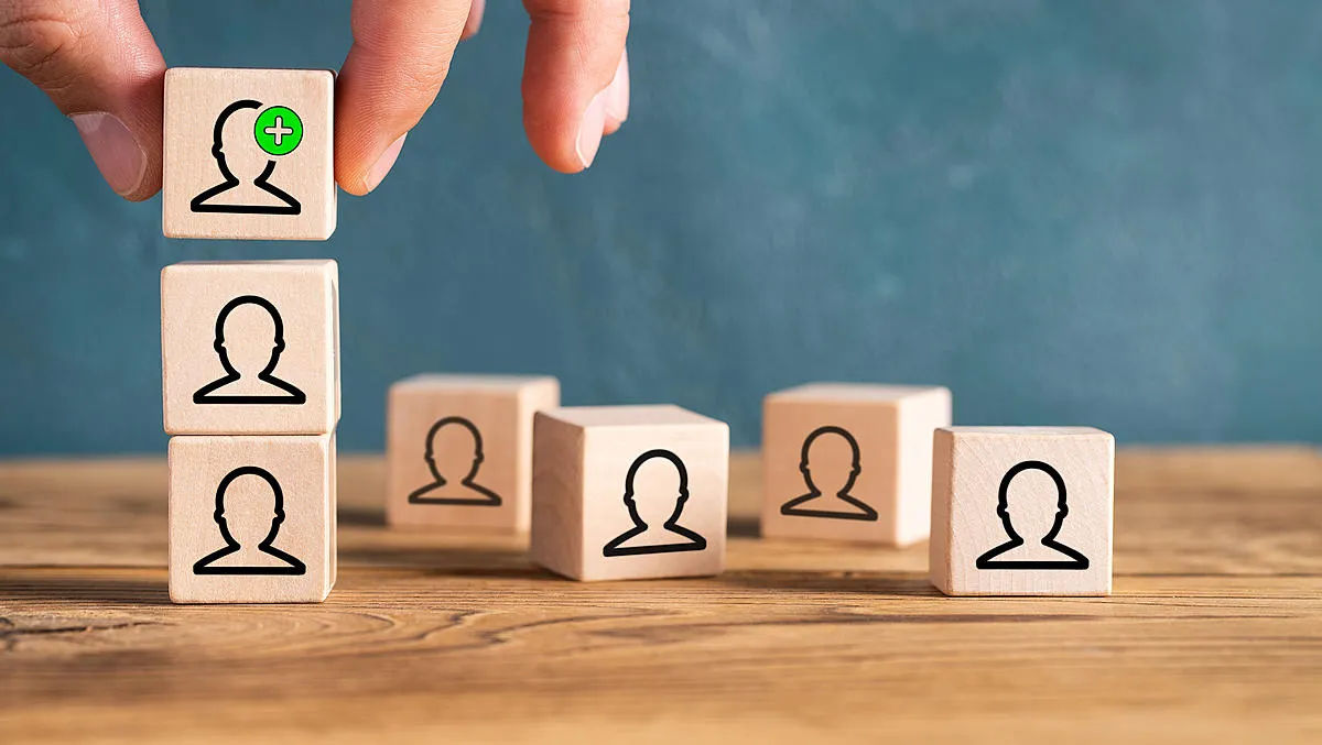 Hand stacking wooden blocks with user profile icons, one block with a green plus sign indicating adding a new person, on a wooden surface with a blue-gray background.
