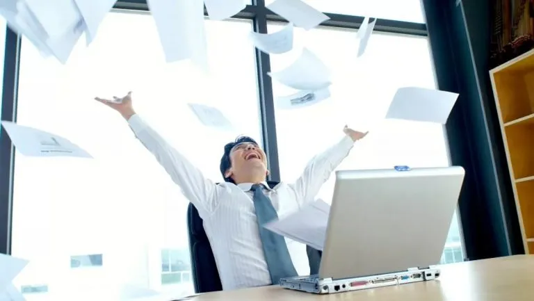 Excited man in a shirt and tie sitting at a desk with a laptop, throwing papers in the air in an office with large windows.
