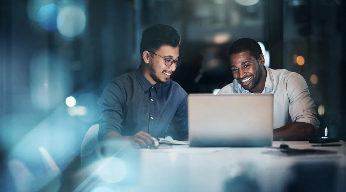 Two men sitting at a table working together on a laptop, both smiling in a dimly lit office environment.