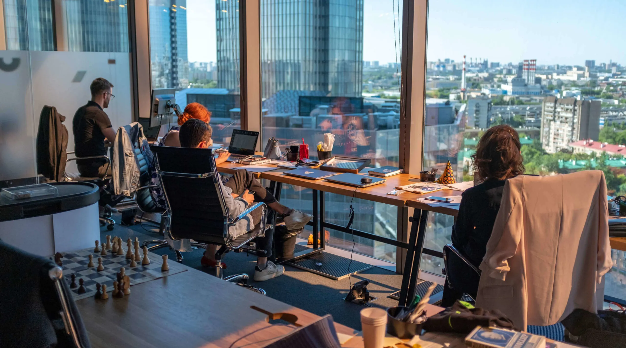 Four people working at desks in a modern office with large windows overlooking a cityscape of tall buildings and residential blocks during daytime.