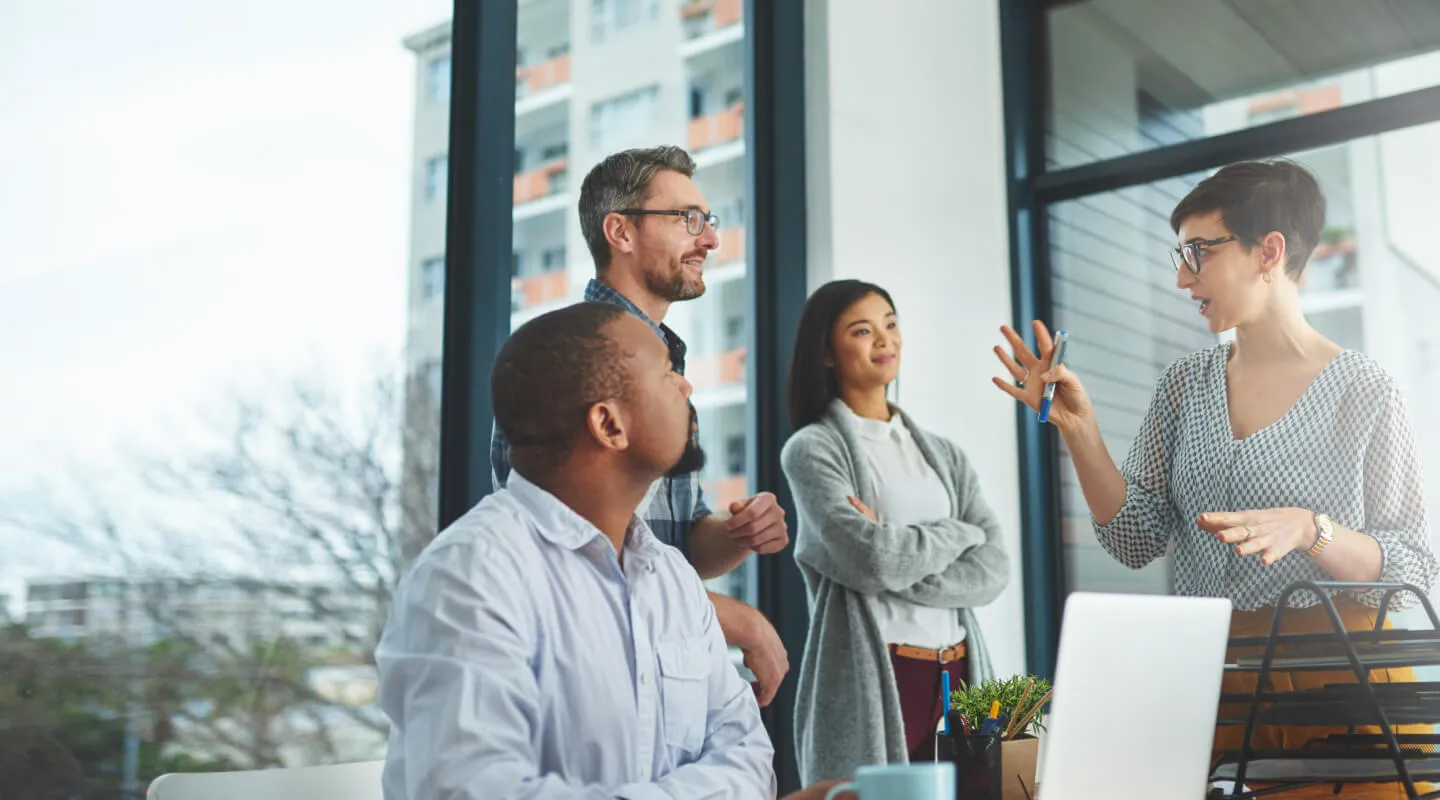 Four colleagues engaged in a discussion in a modern office with large windows, one woman speaking and gesturing with a pen while the others listen attentively.