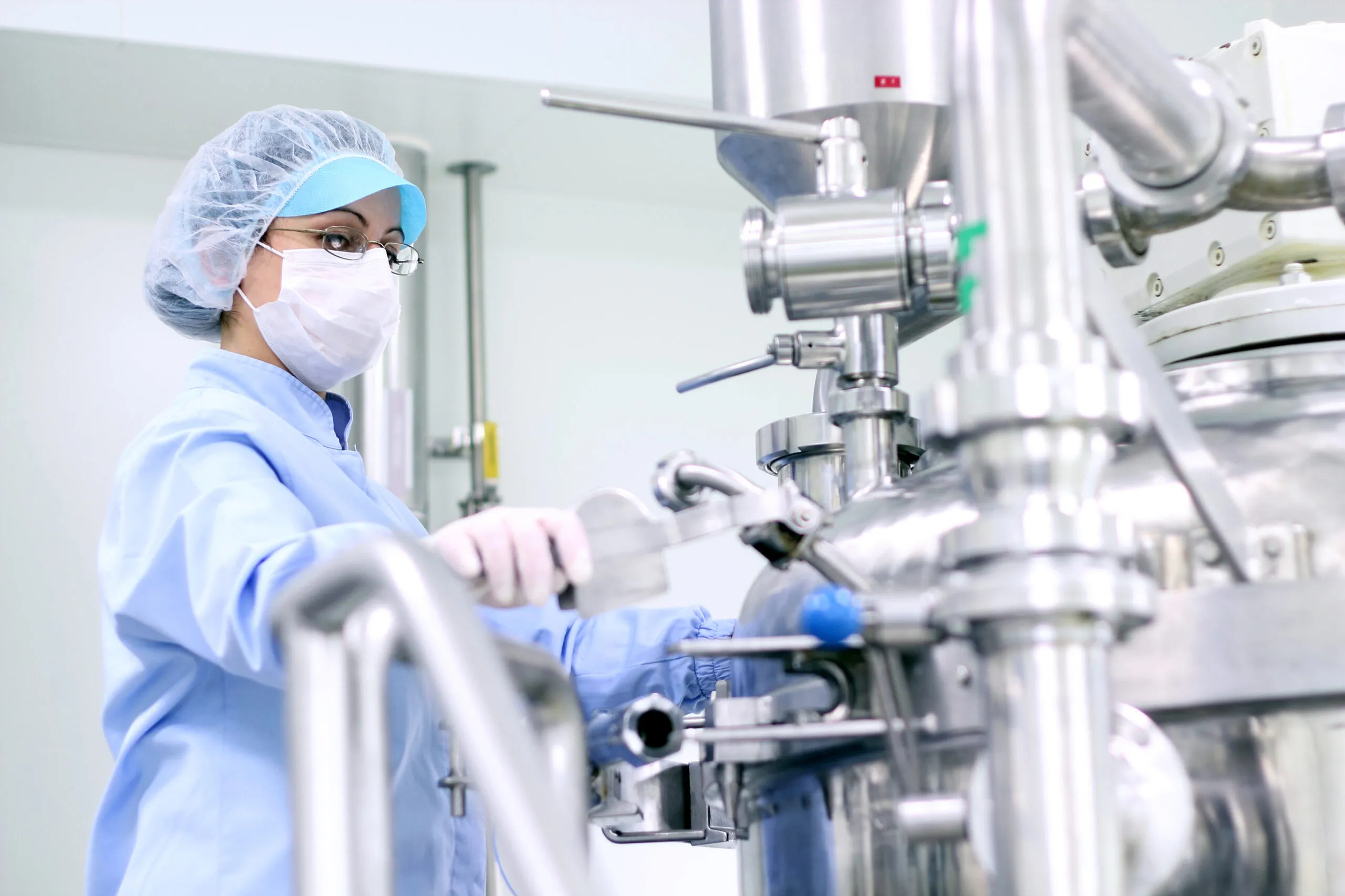 Female laboratory technician wearing a blue protective suit, hairnet, mask, and glasses, operating stainless steel pharmaceutical or chemical processing equipment in a clean room.