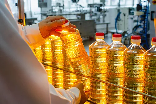 Person inspecting a clear plastic bottle filled with golden liquid, surrounded by similar bottles in an industrial setting.