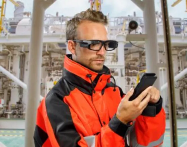 Man wearing an orange and black safety jacket and augmented reality glasses looking at a handheld device, with an industrial facility background.