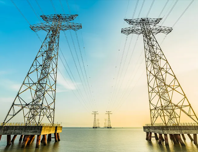 Four large electrical transmission towers standing in a body of water, with wires stretching between them under a blue and light orange sky.