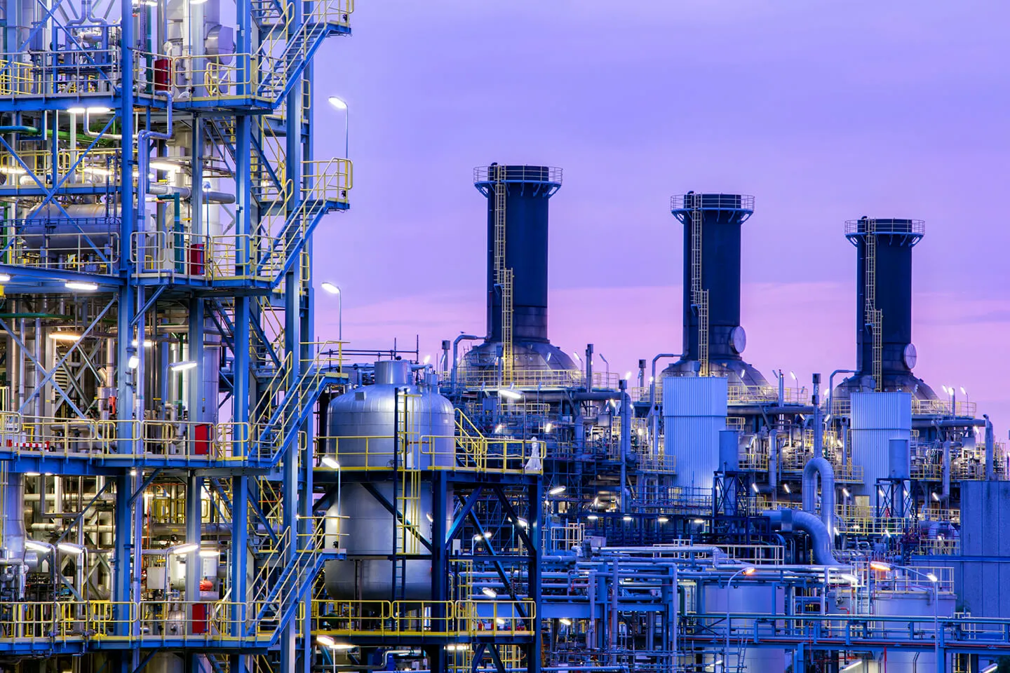 Industrial chemical plant with complex metal structures, tanks, pipes, and large exhaust stacks against a twilight sky with a purple hue.