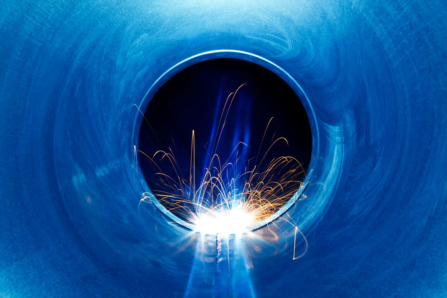 Inside view of a blue metal pipe with bright welding sparks and light at the bottom center.
