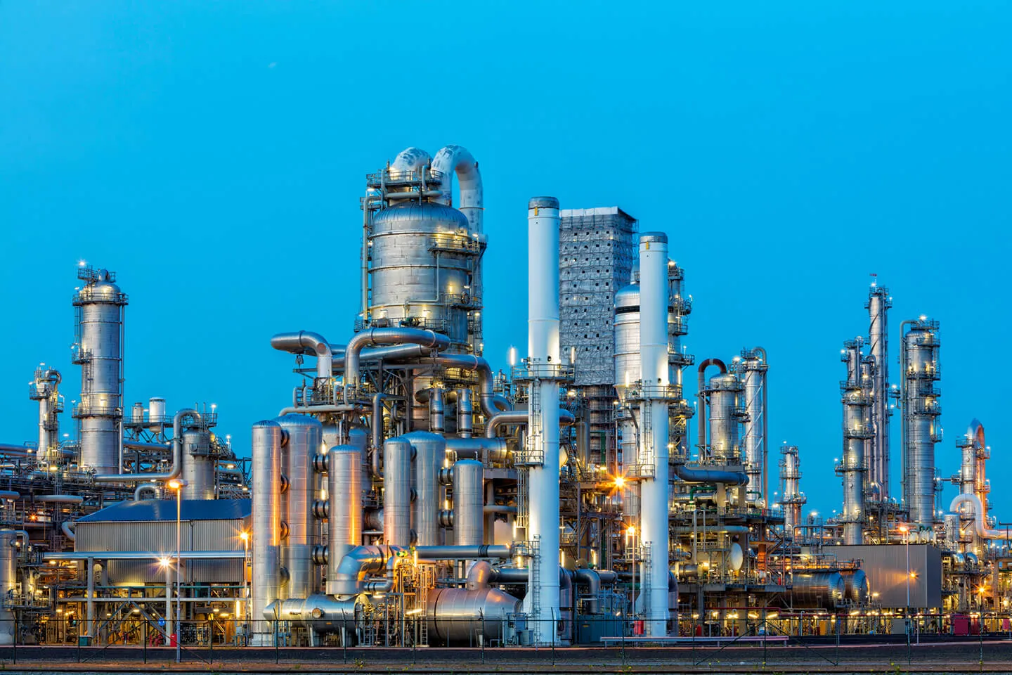 Large industrial oil refinery complex with numerous metallic distillation towers, pipelines, and processing equipment illuminated against a clear blue sky at dusk.