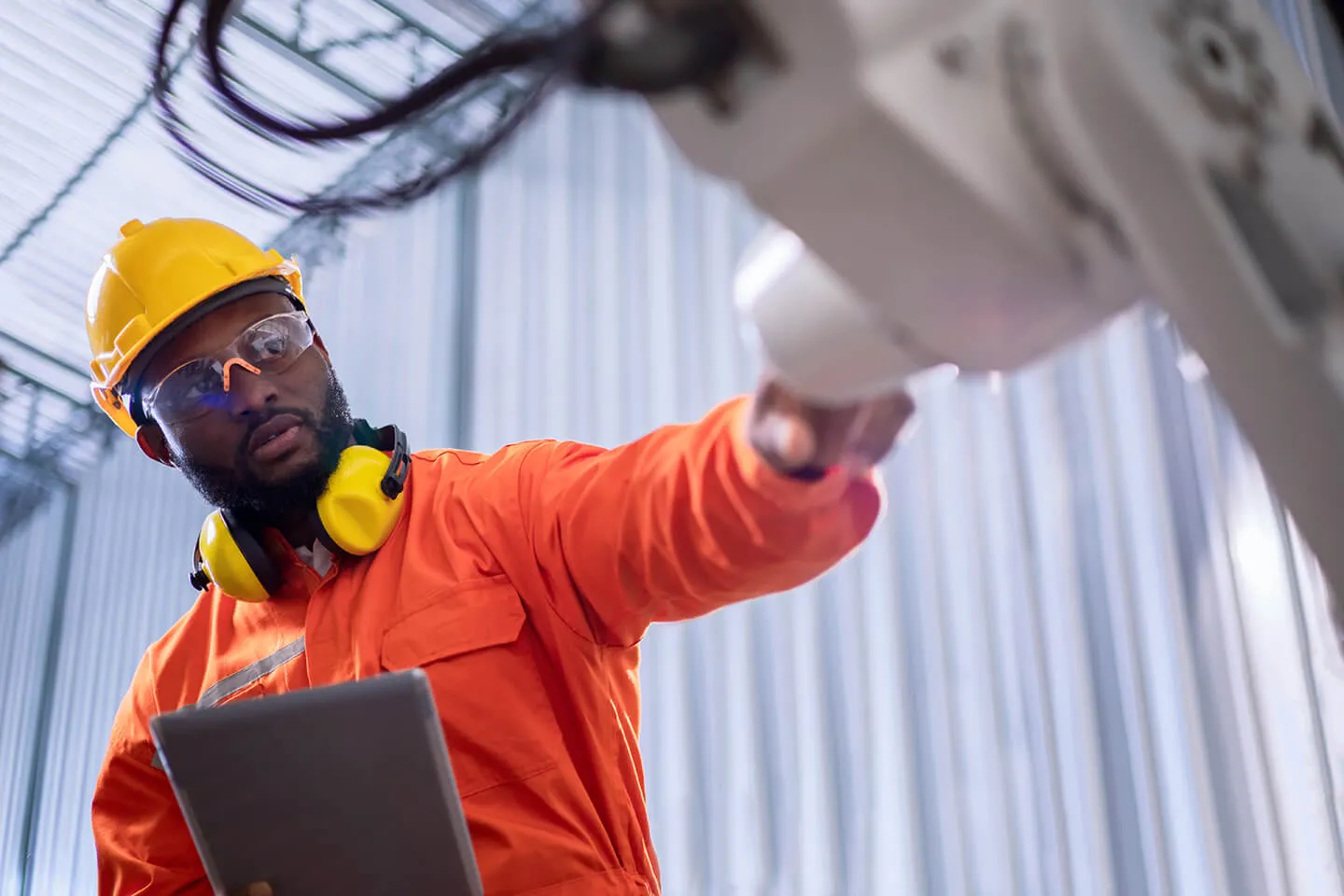 Man in orange safety coveralls, yellow hard hat, and protective glasses holding a tablet and pointing at industrial equipment inside a facility.