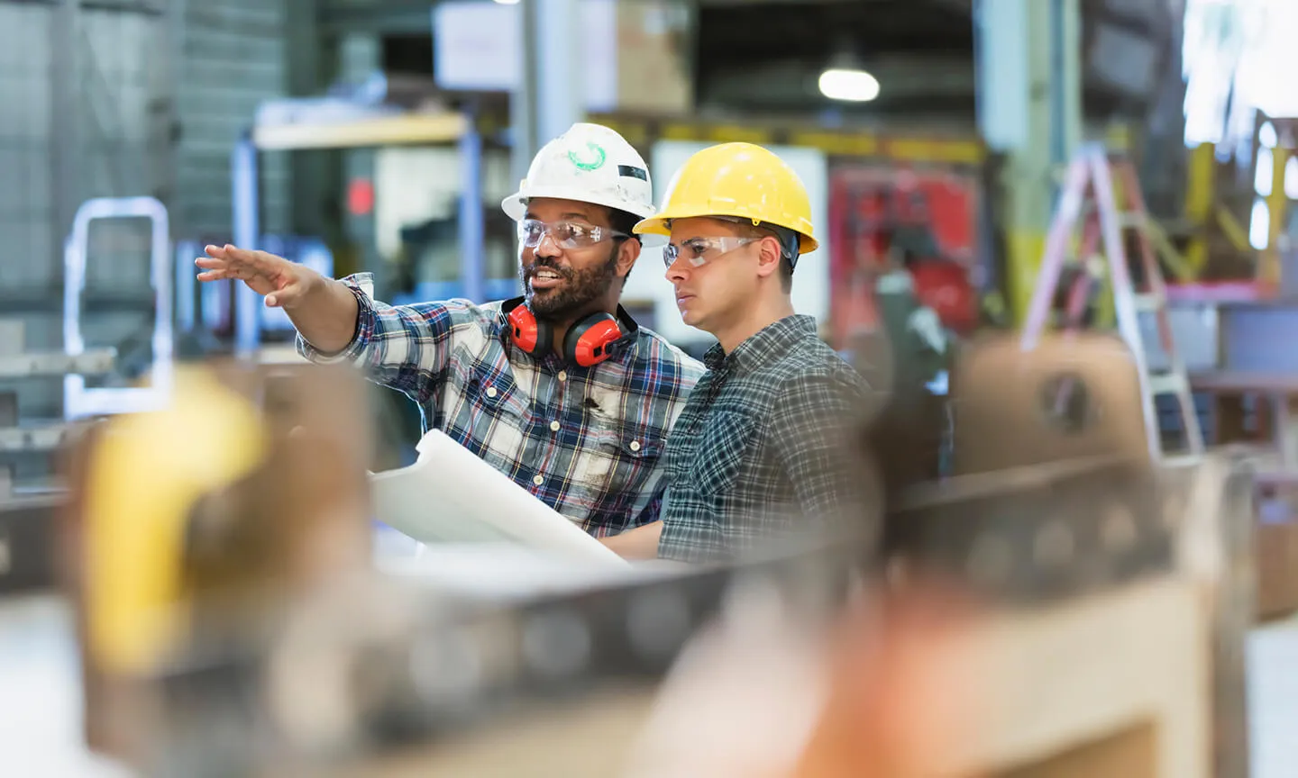 Two male industrial workers wearing hard hats and safety glasses discuss plans inside a factory, with one pointing ahead while holding blueprints.