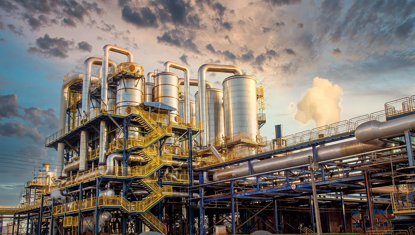 Industrial facility with multiple large cylindrical metal tanks and interconnected silver pipes against a dramatic cloudy sky at sunset, featuring yellow staircases and railings.