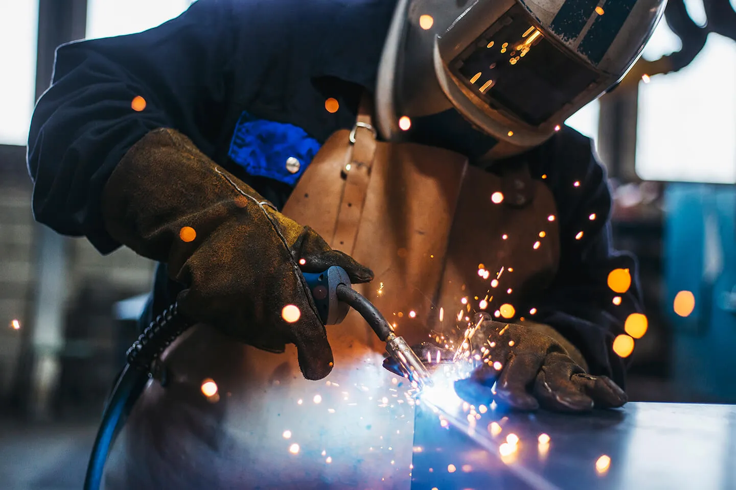Worker in protective gear welding metal, with sparks flying from the welding torch in an industrial setting.