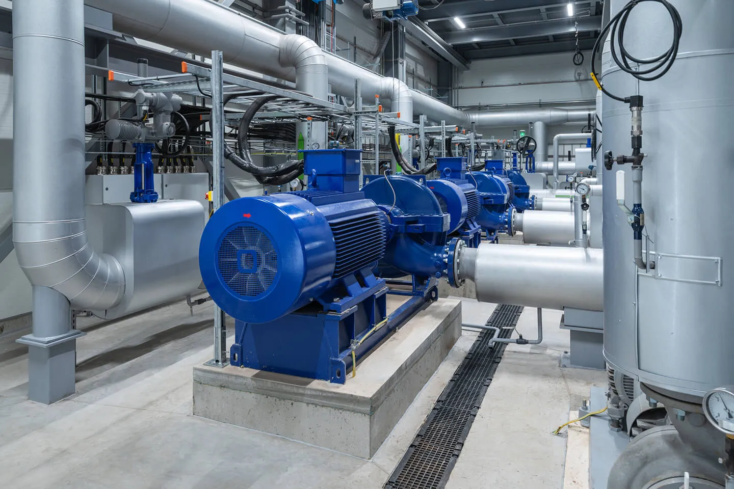 Row of large blue industrial electric motors connected to silver piping inside a clean, modern mechanical room with concrete floor and metal ductwork.