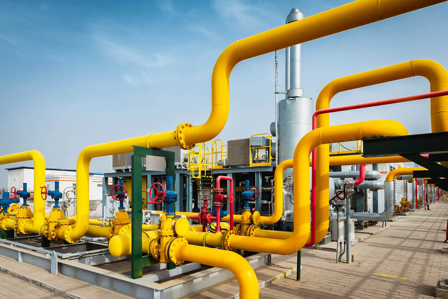 Industrial setup with a network of large yellow pipes and valves with red and blue handles under a clear blue sky, adjacent to metal structures and equipment on a paved ground.