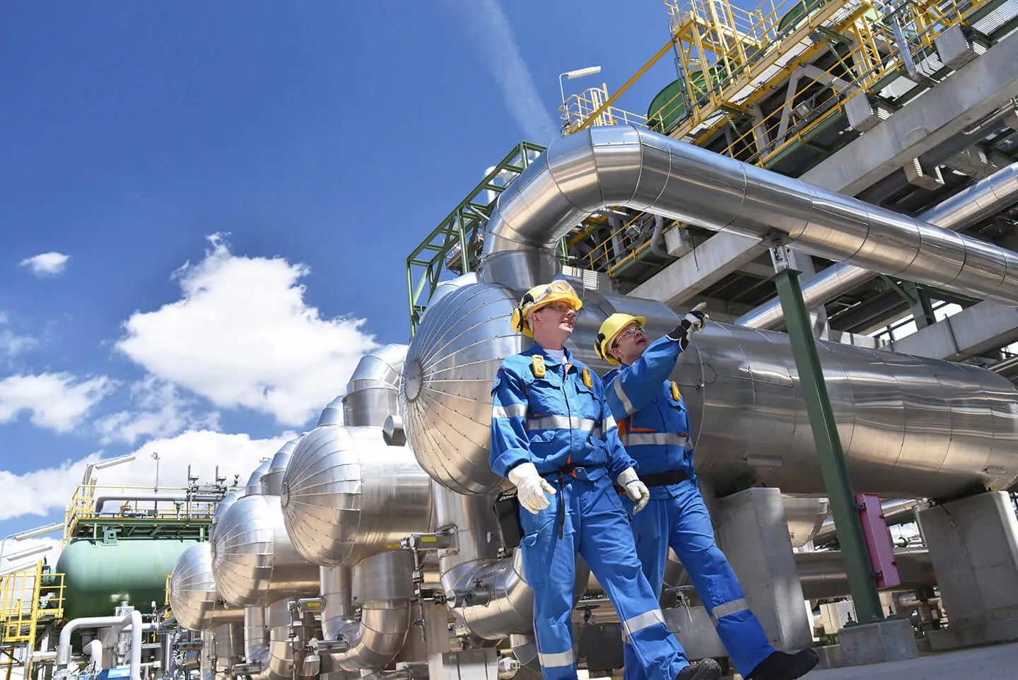 Two industrial workers wearing blue protective uniforms and yellow hard hats stand beside large metallic pipes and tanks at a refinery or processing plant under a bright blue sky with clouds. One worker points towards the equipment while the other observes.