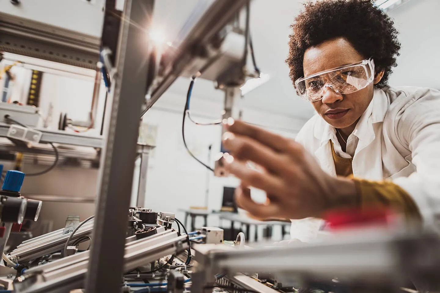 A woman wearing safety goggles and a lab coat working on a complex machine with wires and metal components in a laboratory or industrial setting.
