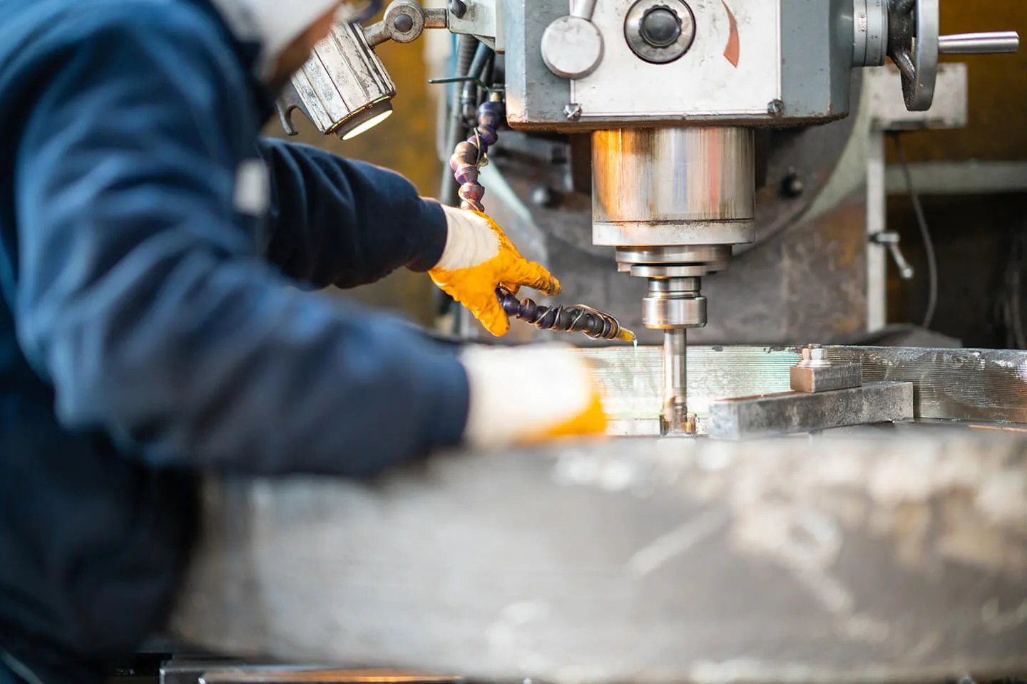 Worker in orange gloves operating a metal milling machine, machining a large metal piece with coolant fluid applied.