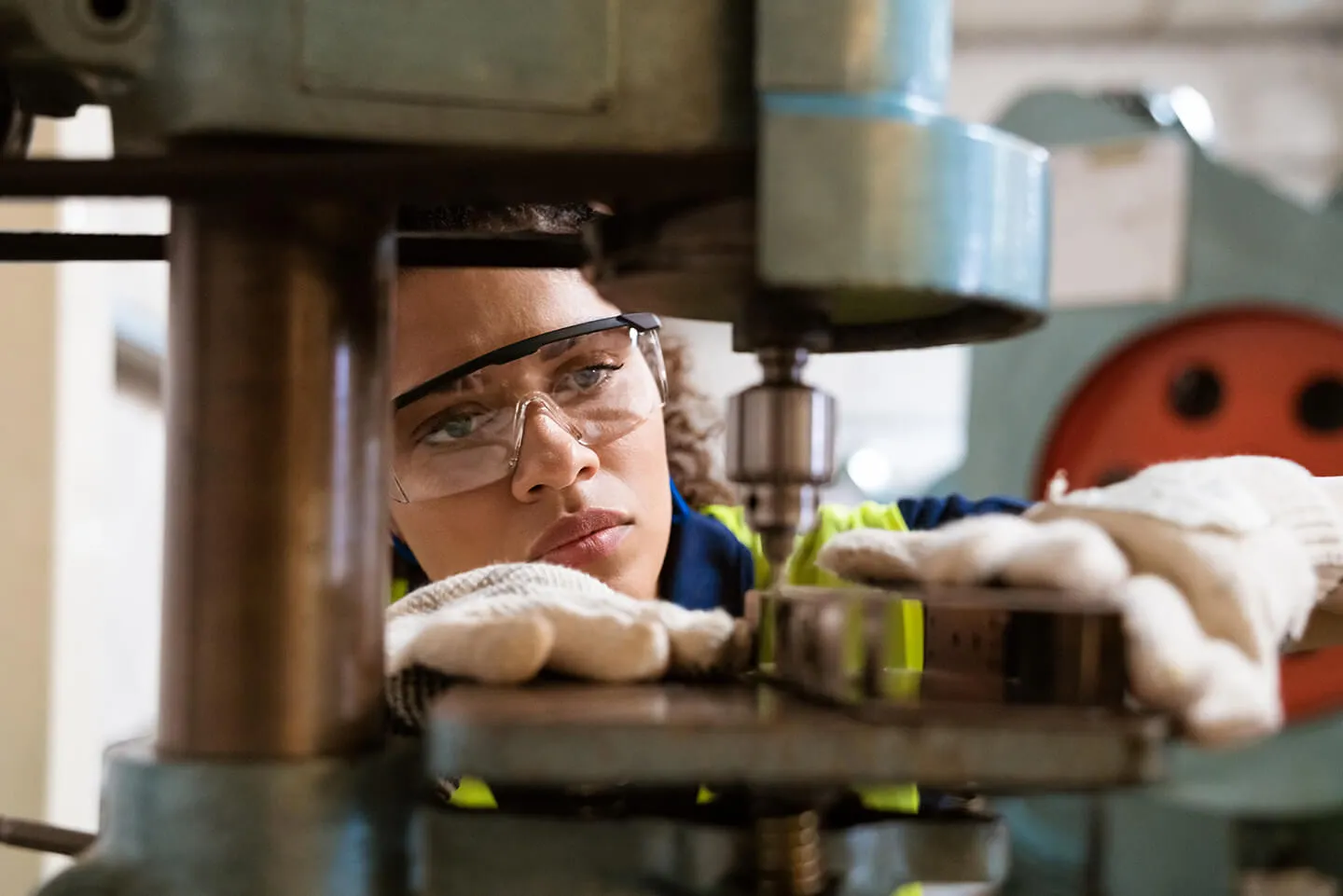 Woman wearing safety goggles and gloves intently operating a metalworking machine in an industrial setting.