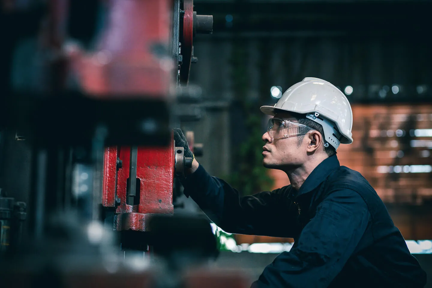 Industrial worker wearing a white hard hat and safety glasses operates a large red machine in a factory setting with blurred background elements.