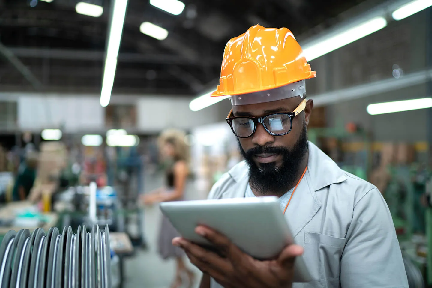 Male factory worker wearing an orange hard hat and glasses using a digital tablet in an industrial setting with machinery and blurred background workers.