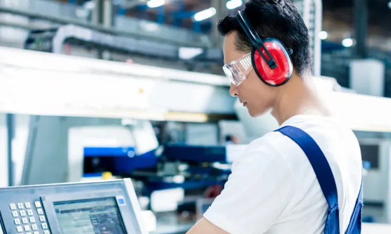 Man wearing red ear protection and safety glasses works on a computer in a factory or industrial setting.