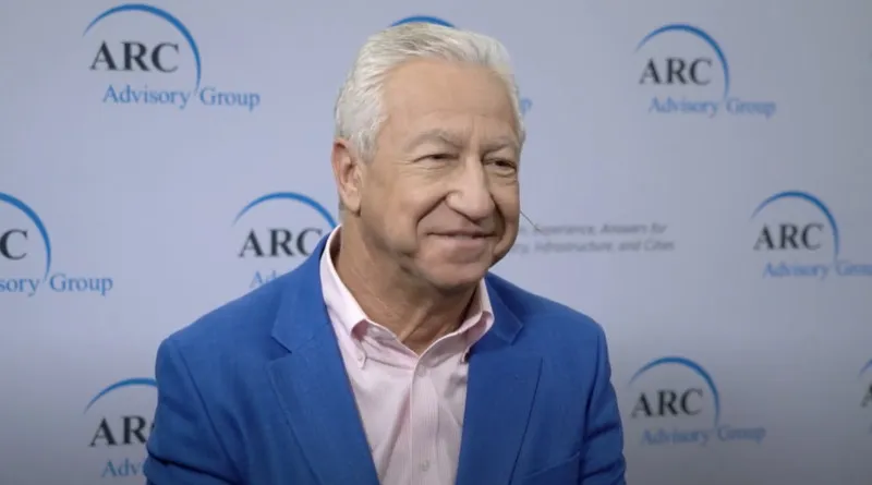 Smiling older man with short white hair wearing a blue blazer and light pink shirt, standing against a backdrop displaying the ARC Advisory Group logo.