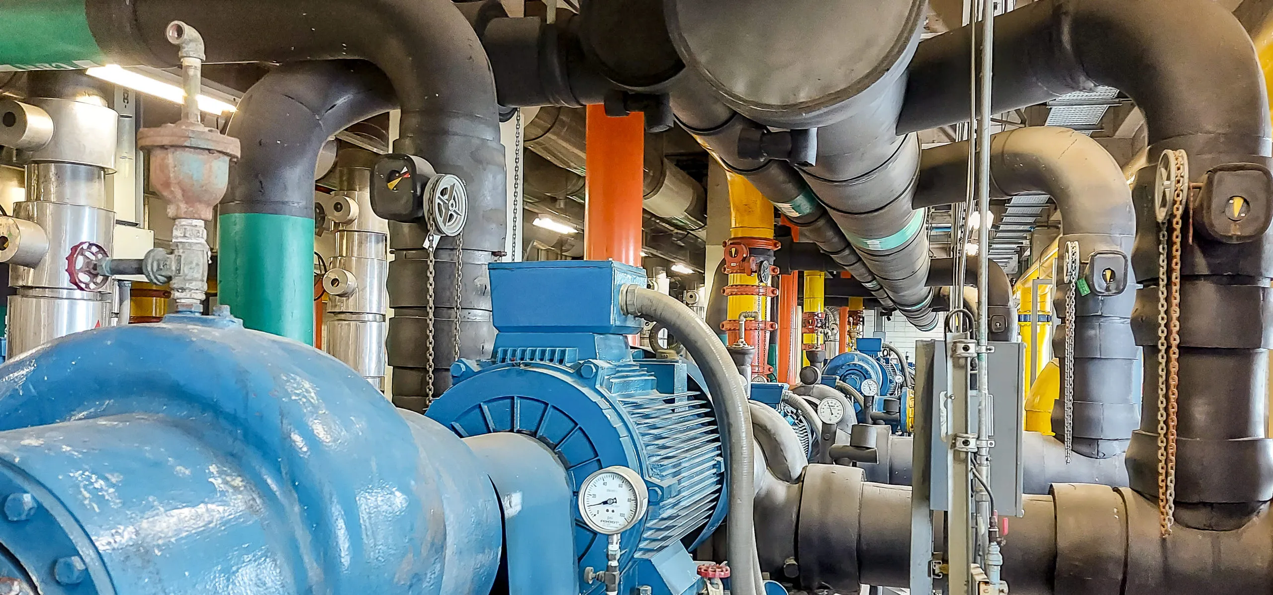 Industrial room with large blue motors and extensive network of insulated black pipes with pressure gauges and valves, supported by bright orange and yellow structural columns.