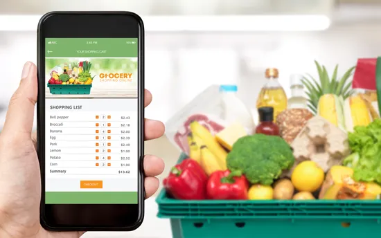 Hand holding smartphone displaying an online grocery shopping cart with items and prices; a green basket of fresh groceries including broccoli, bell peppers, bananas, pineapple, and corn is in the background.