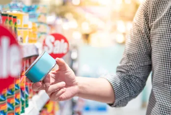 Person in checkered shirt holding a blue canned product near a grocery store shelf with red sale signs.