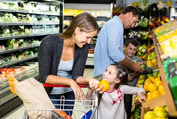 Mother and young daughter smiling while selecting an orange from a fruit display in a grocery store, with a man and child in the background picking produce near refrigerated shelves.