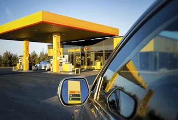 View from a car driver's side mirror showing a gas station with yellow and red canopy and fuel pumps, a convenience store in the background, and another car parked nearby.