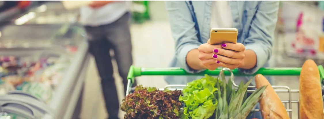 Person with purple nail polish holding a smartphone while pushing a grocery cart filled with fresh vegetables and baguettes in a supermarket aisle.