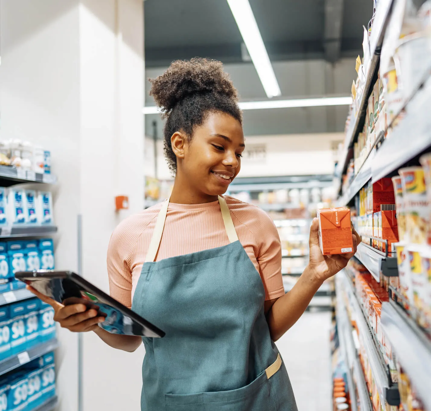 A smiling woman in a store aisle wearing an apron holds a tablet in one hand and an orange carton in the other while checking product details.