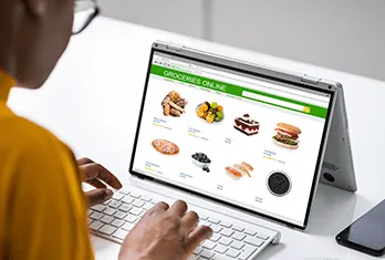 Person using a laptop displaying an online grocery store webpage with images of various food items including bread, vegetables, cake, burger, pizza, blueberries, sushi, and a cookie.