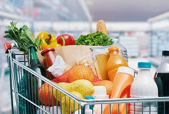 Shopping cart filled with groceries including fresh vegetables, fruits, bread, packaged juice, and milk bottles in a supermarket aisle.