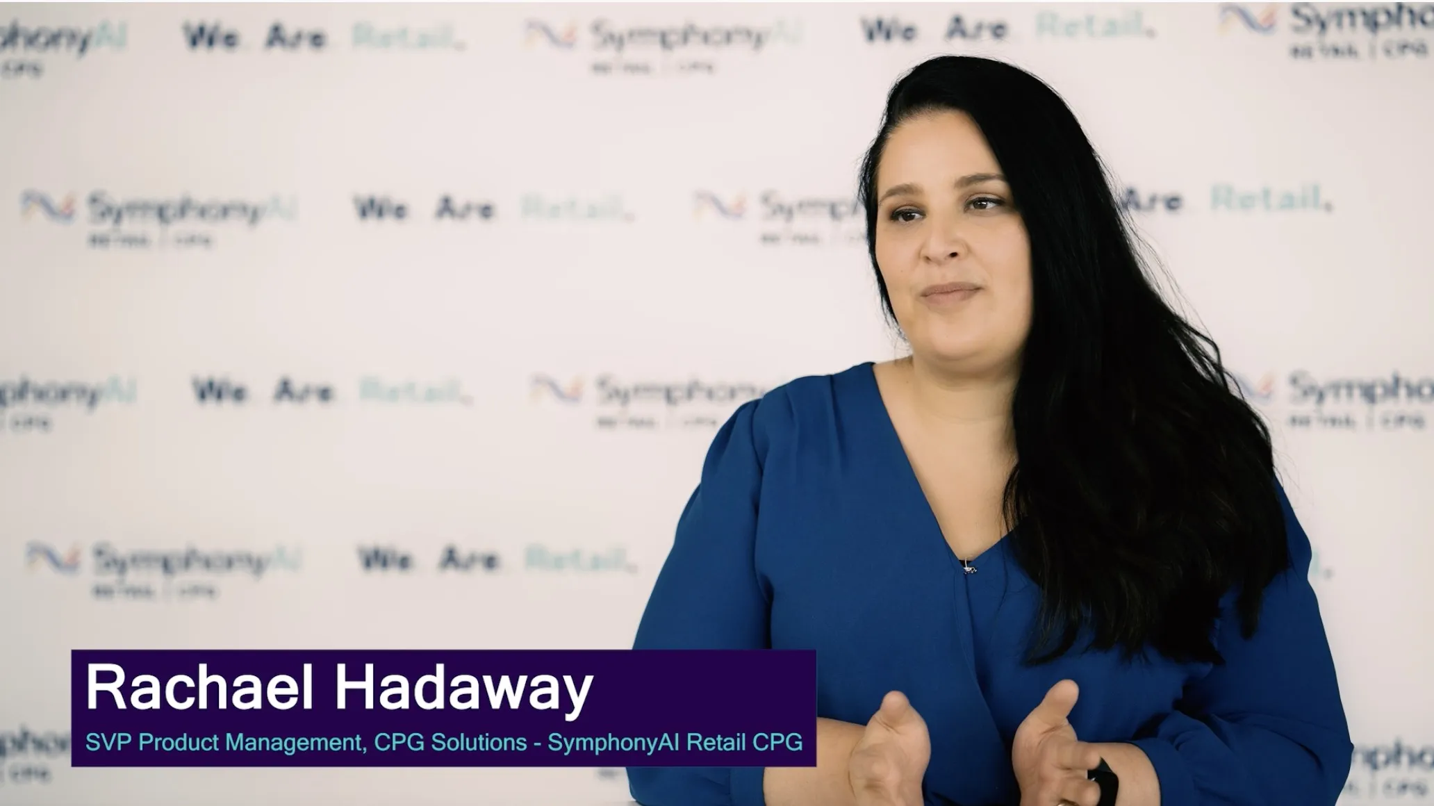 A woman with long dark hair wearing a blue top stands in front of a backdrop with repeated logos and text saying 'We Are Retail' and 'SymphonyAI Retail CPG', with a caption identifying her as Rachael Hadaway, SVP Product Management, CPG Solutions at SymphonyAI Retail CPG.