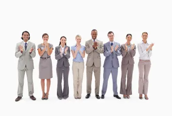 A diverse group of seven professionally dressed people standing in a line, smiling and clapping their hands against a white background.