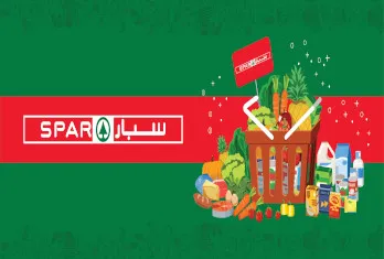 Shopping basket filled with vegetables and groceries including bread, carrots, and corn, alongside various packaged products and dairy containers, with a Spar logo featuring Arabic text on a red and green background.