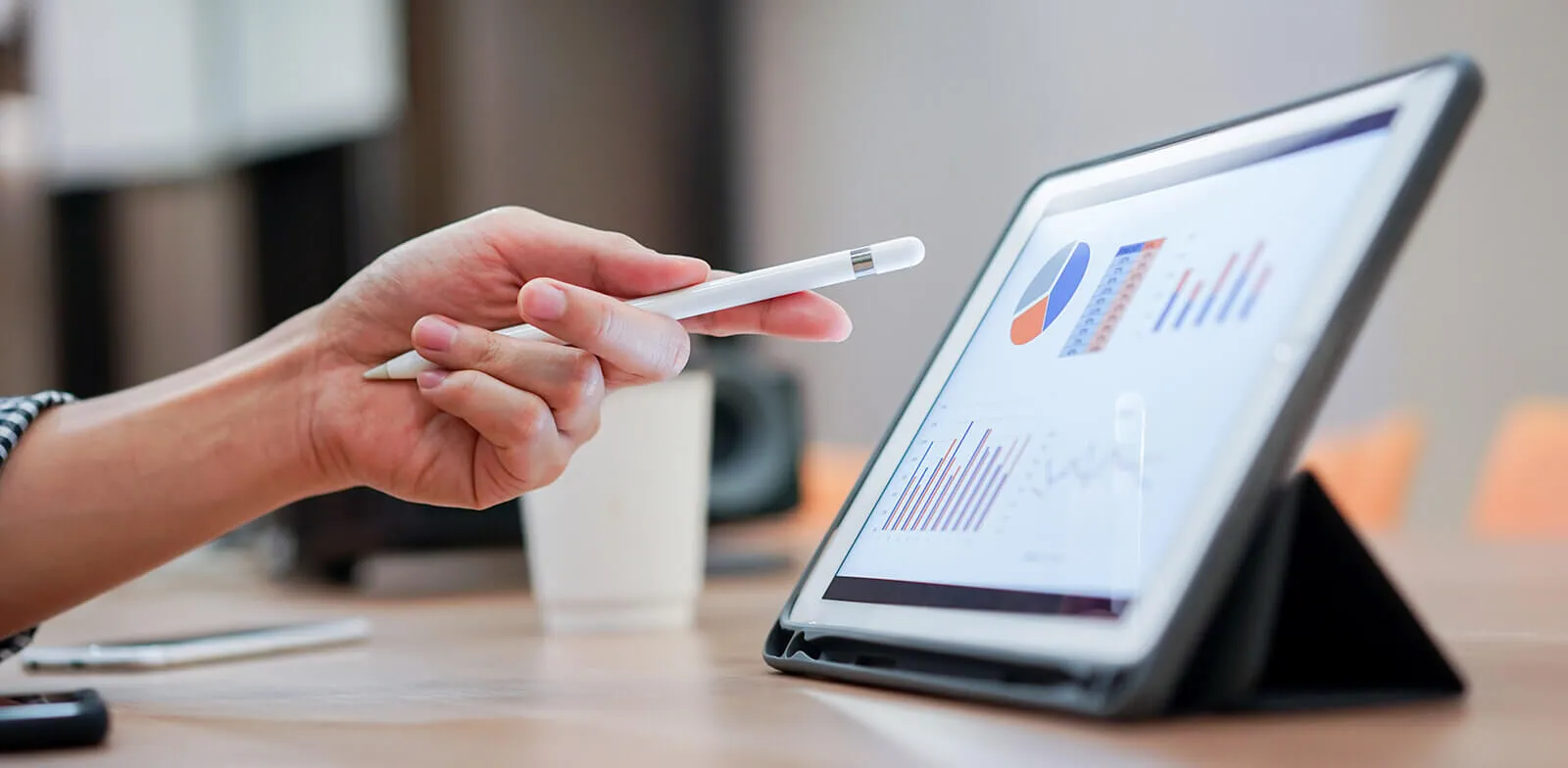 Person holding white stylus pen and pointing at a tablet screen displaying colorful charts and graphs on a desk.