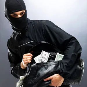 Person in black mask and clothing holding a black bag filled with US dollar bills, looking to the side cautiously.