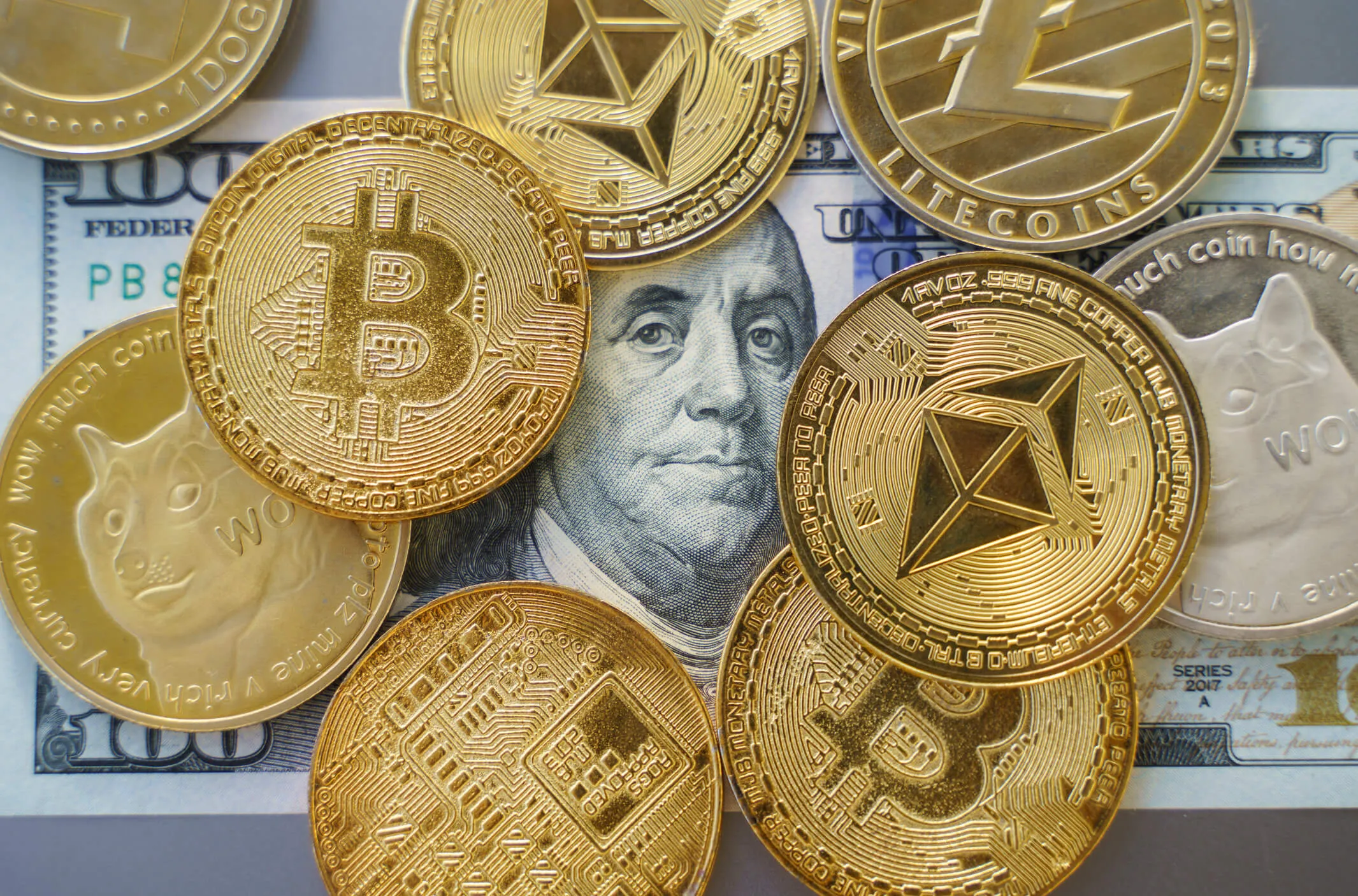 Several physical cryptocurrency coins including Bitcoin, Ethereum, Litecoin, and Dogecoin placed on a US hundred-dollar bill featuring Benjamin Franklin.