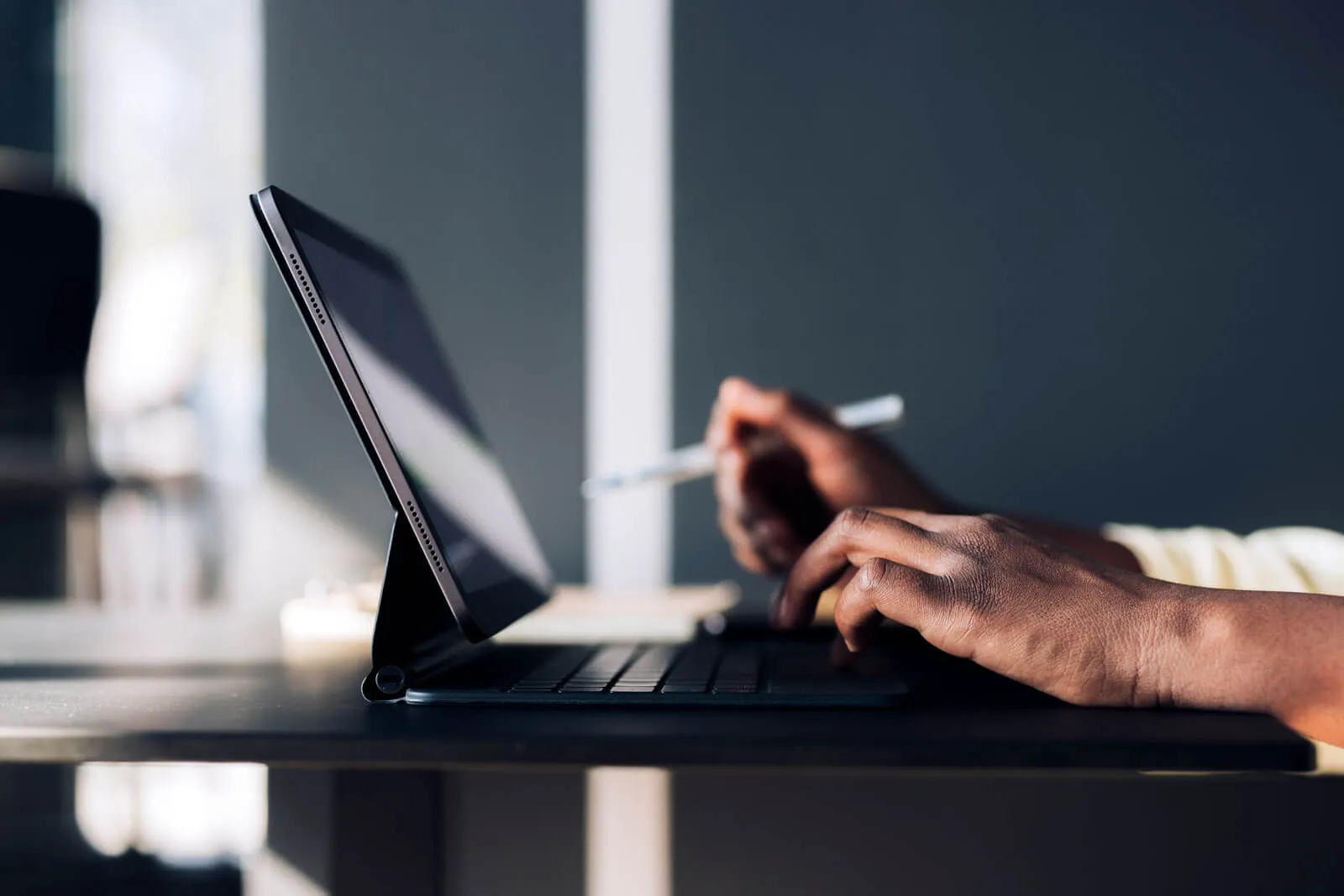 Close-up of a person using a tablet with a keyboard case, holding a stylus pen in one hand and typing on the keyboard with the other.