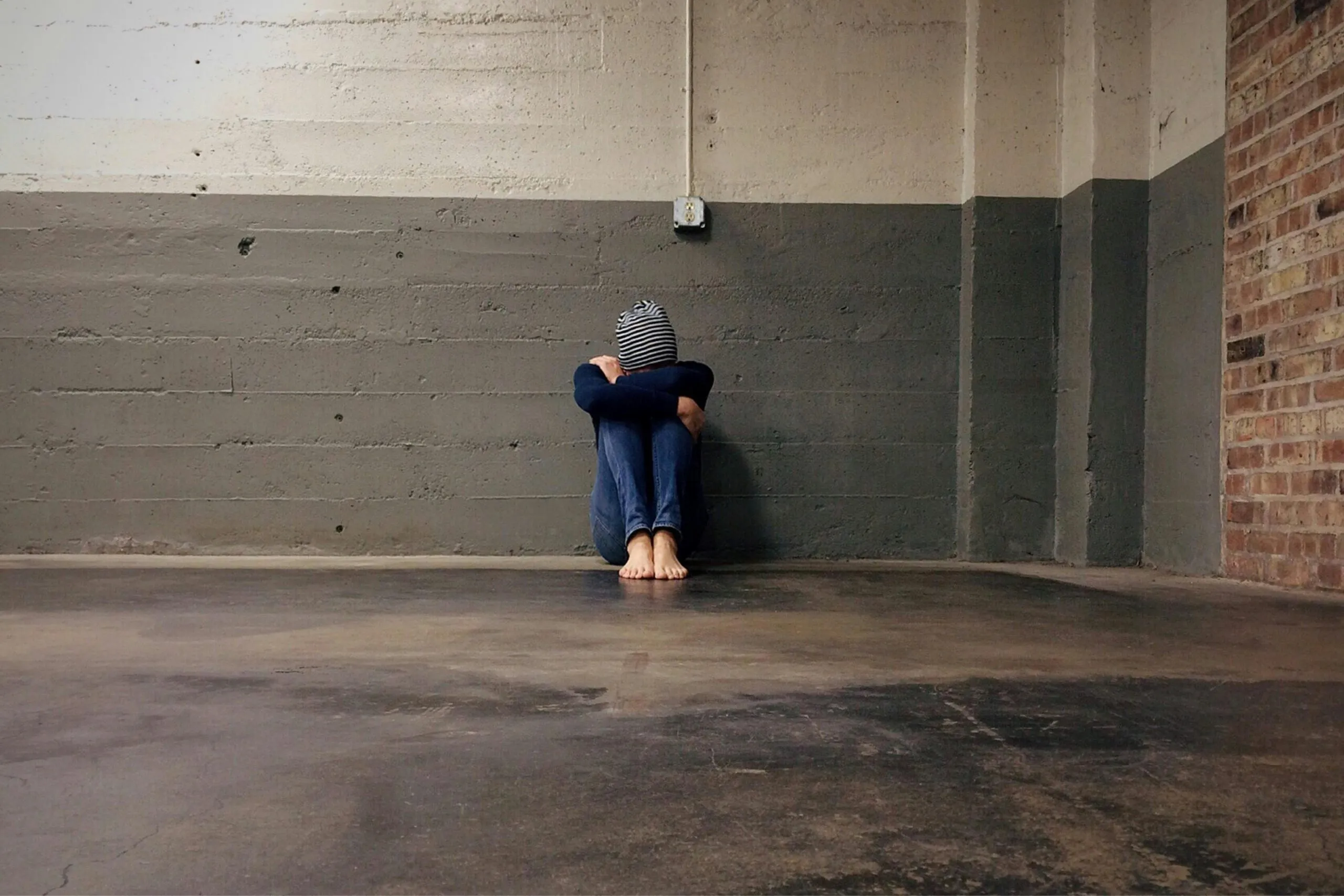 Person sitting barefoot on a concrete floor in a corner with knees hugged to the chest and head covered by a striped cloth, suggesting distress or isolation.