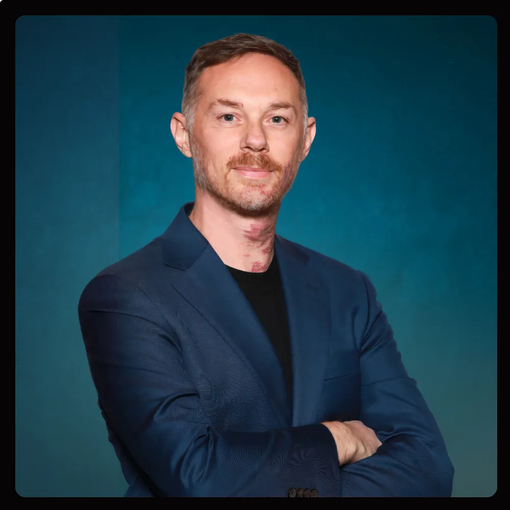Portrait of a man with light skin, short brown hair, and a trimmed beard, wearing a dark blue blazer over a black shirt, standing with arms crossed against a blue background.