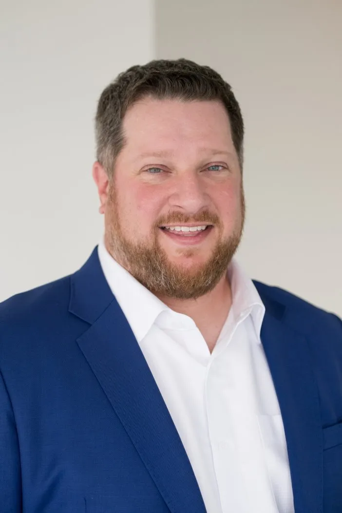 Portrait of a smiling man with short brown hair and a beard wearing a white shirt and blue suit jacket against a plain light background.