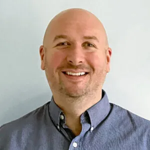 Smiling bald man with light skin wearing a buttoned gray shirt against a plain light background.