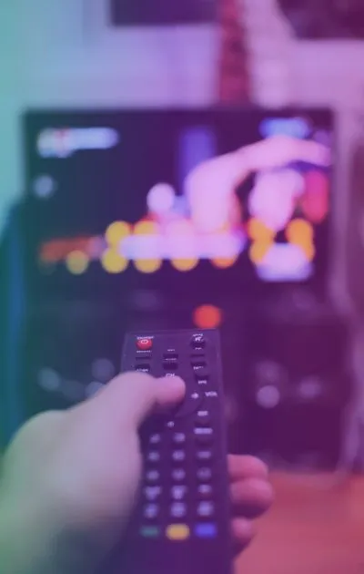 Hand holding a TV remote pointed toward a television screen displaying blurred colorful graphics.