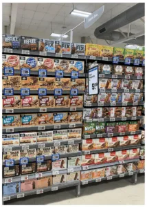 Grocery store aisle shelf displaying various types of packaged breakfast cereals and snack bars arranged in rows with price tags below each item.