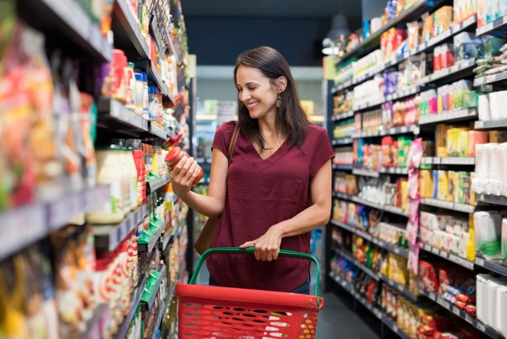 Smiling woman in a maroon shirt holding a red shopping basket with one hand and examining a product jar from a grocery store shelf.