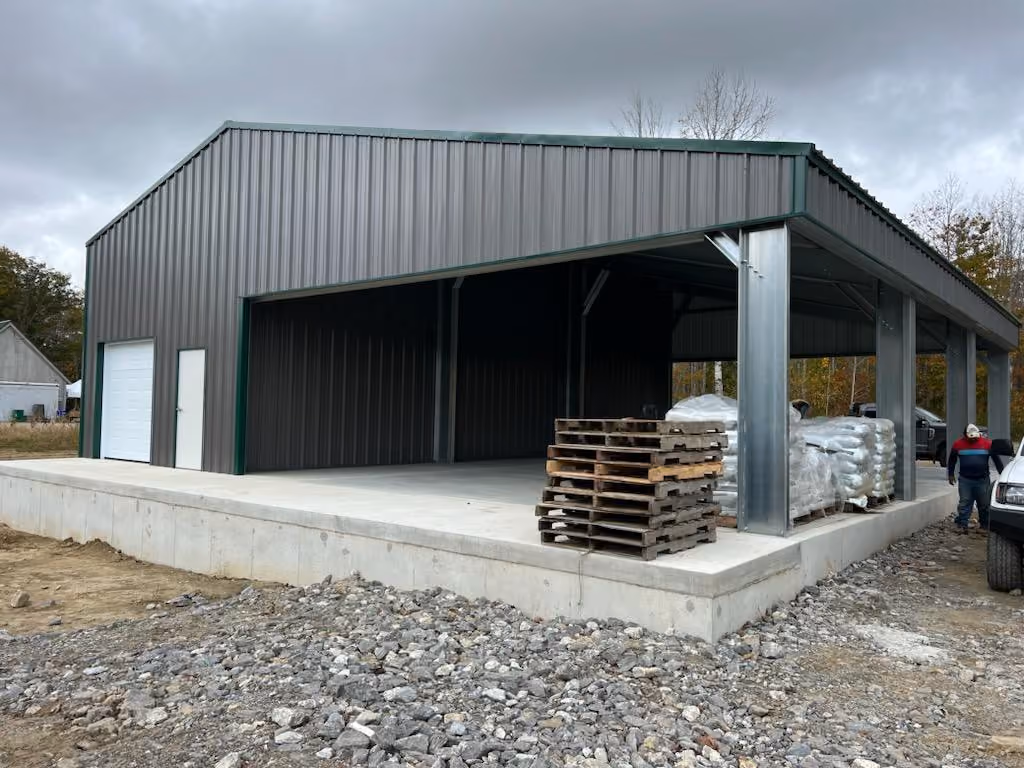 Metal building with a large open side, a closed section with a door and roll-up door, pallets stacked on a concrete foundation, under a cloudy sky.